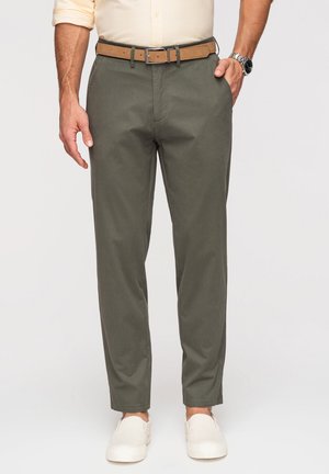 Homme portant un pantalon décontracté vert olive, une ceinture marron clair, une chemise beige aux manches retroussées, des chaussures blanches sans lacets et une montre-bracelet au poignet gauche.