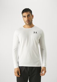 Bijela košulja dugih rukava izrađena od glatke tkanine, s okruglim izrezom i crnim Under Armour logotipom na gornjem lijevom dijelu prsnog koša.