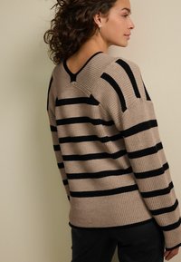 Beige gestreifter Pullover mit schwarzen horizontalen Linien, gerippter Textur, V-Ausschnitt hinten und enganliegenden Ärmeln, kombiniert mit schwarzen Hosen.
