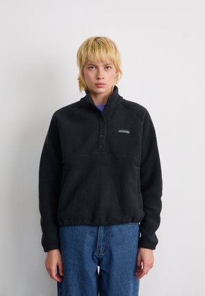 Ungdom med kort blondt hår iført en svart Columbia fleecejakke og blå jeans, stående mot en enkel hvit bakgrunn.