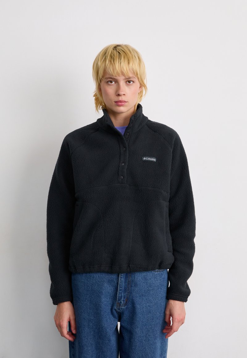 Giovane con capelli biondi corti che indossa una giacca in pile nera Columbia e jeans blu, in piedi contro uno sfondo bianco uniforme.