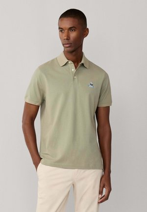 Hombre con polo de manga corta color verde claro con pequeño logo bordado, pantalones beige, de pie con una mano en el bolsillo contra fondo liso.