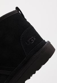 Stivale alla caviglia in suede nero con suola in gomma piatta, dotato di un tassello per il tiro e dettaglio del logo incastonato sul lato. La parte superiore ha una texture liscia.