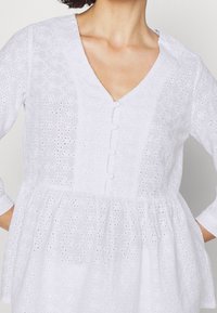 Weiße Broderie anglaise Bluse mit 3/4-Ärmeln, V-Ausschnitt, mit Stoff überzogenen Knöpfen und gerafftem Peplum-Taille, getragen von einer Person.