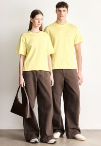 Żółte bawełniane t-shirty z krótkimi rękawami i diamentowymi ozdobami, połączone z oversize'owymi brązowymi spodniami. Brązowa torebka dopełnia stylizację.