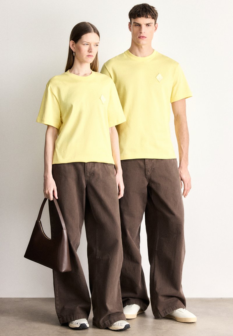 Magliette di cotone gialle con maniche corte e dettagli a diamante, abbinate a pantaloni marroni oversize. Una borsa marrone completa il look.