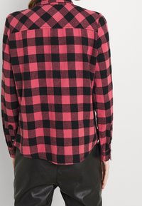 Checkered shirt in pink and black flannel, featuring a button-up front, long sleeves, and a rounded hem. Texture appears soft.