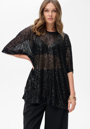 Vrouw in een doorschijnende zwart pailletten top over een zwarte beha met zwarte broek, posing met één hand op de heup tegen een witte achtergrond.