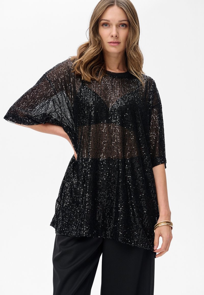 Kobieta w przezroczystej, czarnej bluzce z cekinami, nosząca czarny stanik i czarne spodnie, pozująca z jedną ręką na biodrze na białym tle.