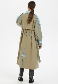 Kvinna med långt hår i en beige trenchcoat med ljusblå detaljer, grå randiga byxor och svarta skor, stående vänd bort.