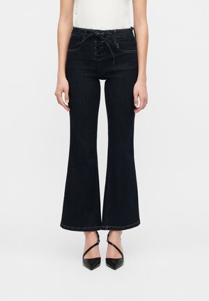 Frontalt billede af en person iført højttaljede sorte flared jeans med snøredetalje og sorte spidse hæle med krydsede stropper.