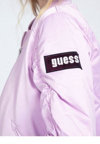 Giacca bomber di colore viola chiaro con chiusura a zip, colletto a costine e un patch ricamato "guess" in bianco e nero sulla manica sinistra.