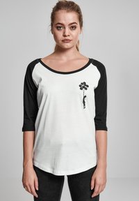 Zwart-wit honkbal T-shirt met driekwartmouwen. Voorkant heeft een afbeelding van een figuur die ballonnen vasthoudt. Zacht materiaal, losse pasvorm.