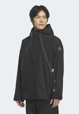 Mlad moški, oblečen v črno kapuco Adidas dežni plašč z odsevnimi detajli, ki gleda levo proti svetlemu ozadju.