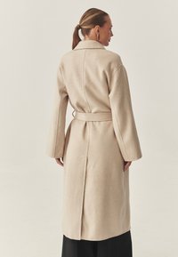 Bež plašč iz mešanice volne z pasom na pasu, prevelikimi rokavi in ovratnikom. Ima sredinski zadnji šiv in razporek na spodnjem robu.