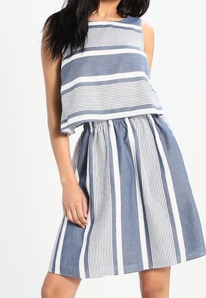 Frau trägt ein ärmelloses, blau-weiß gestreiftes bauchfreies Top und einen passenden hoch taillierten Rock, steht mit einer Hand entspannt.