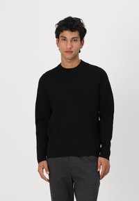 Schwarzer Strickpullover mit Rundhalsausschnitt, langen Ärmeln und glatter Textur. Getragen mit grauen Hosen, vor einem einfarbigen weißen Hintergrund gezeigt.