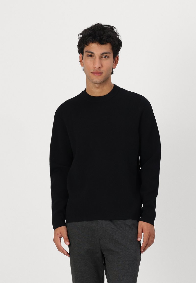 Schwarzer Strickpullover mit Rundhalsausschnitt, langen Ärmeln und glatter Textur. Getragen mit grauen Hosen, vor einem einfarbigen weißen Hintergrund gezeigt.