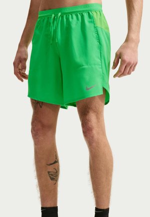 Mand iført lysegrønne Nike sportsshorts med sidelommer og snøre i taljen, der står med synlige tatoveringer på benene.