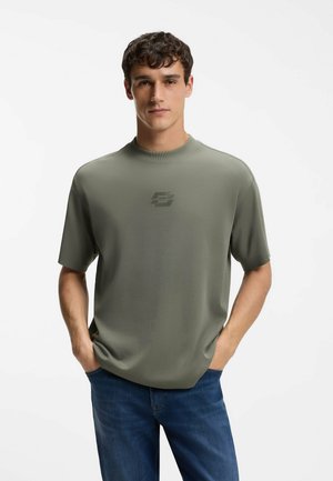 Mand iført olivengrøn kortærmet skjorte med subtilt logo og blå jeans, stående med hænderne i lommerne mod hvid baggrund.