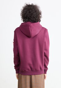Persona con cabello rizado vista desde atrás, usando una sudadera con capucha color burdeos y una falda de cuadros marrón, contra un fondo blanco liso.
