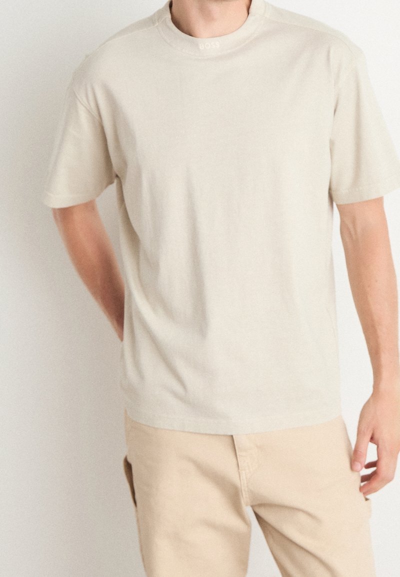 T-shirt beige clair à col rond, fabriqué en coton doux, avec des manches courtes et une coupe relaxée. Présente un logo subtil au niveau de l'encolure.