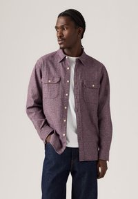Flannel shirt in purple and red hues with a textured pattern, two chest pockets, and cream buttons, paired with dark denim jeans.