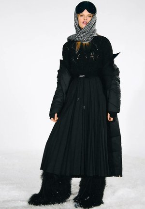 Donna che indossa un outfit invernale nero a strati con lunga gonna plissettata, giacca imbottita, stivali pelosi, sciarpa a quadri e maschera nera per dormire sulla testa.