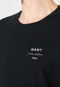 T-shirt en coton noir avec un col rond, arborant un logo brodé blanc et le texte "GANT American Sportswear 1949" sur la poitrine.