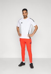Camiseta deportiva blanca con franjas negras en los costados, combinada con pantalones rojos brillantes con toques azules y diseño ajustado. Zapatillas deportivas negras.