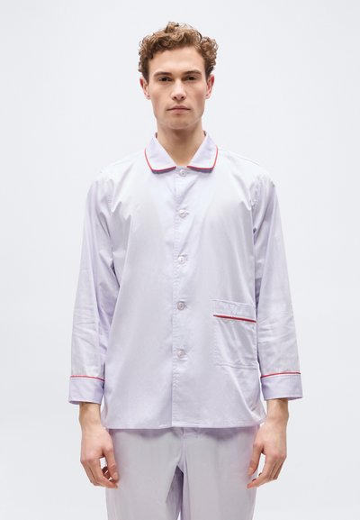 Jeune homme portant un pyjama à manches longues violet clair avec un liseré rouge sur le col, les poignets et la poche, debout devant un fond uni.