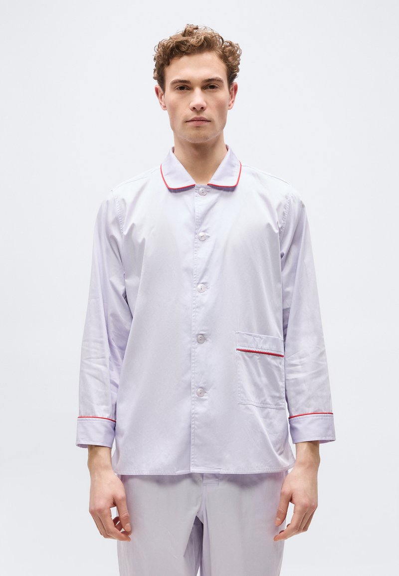 Νέος άνδρας που φοράει μακρυμάνικη πυτζάμα ανοιχτού μωβ χρώματος με κόκκινη ραφή στο γιακά, τις μανσέτες και την τσέπη, στέκεται μπροστά σε απλό φόντο.