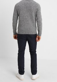 Pull gris tricoté avec un motif texturé, assorti à un pantalon bleu marine et des baskets blanches, vu de dos.