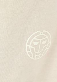T-shirt beige con una trama liscia, caratterizzata da un logo grafico bianco di un volto stilizzato nell'angolo in basso a destra.
