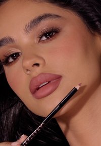 Ołówek do ust z ostrą końcówką, o czarnym korpusie z srebrnym nadrukiem. Modelka prezentuje pełne usta w matowym odcieniu nude.