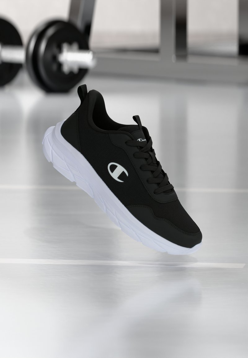 Μαύρο αθλητικό παπούτσι με λευκή σόλα και λογότυπο, να αιωρείται πάνω από το πάτωμα γυμναστηρίου με αλτήρες θολωμένους στο φόντο.