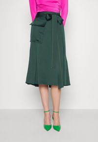 Grön omslagskjol i ett smidigt tyg. Har en stor sidoficka och en rosett-detalj i midjan. Accenterad med kontrasterande sömmar.