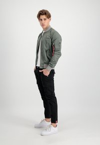 Grön bomberjacka med ribbade muddar och ett bröstpatch, ihopparad med svarta cargobyxor och vita sneakers, som visar en modern avslappnad design.