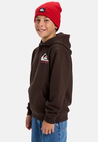 Brown hooded sweatshirt with a front pocket and logo. Paired with blue jeans and a red knit beanie. Features ribbed cuffs and hem.