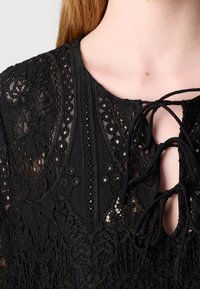 Blouse en dentelle noire avec broderie florale et deux nœuds noués sur la poitrine, montrant une petite ouverture en forme de trou de serrure et une partie du cou et de l'épaule.