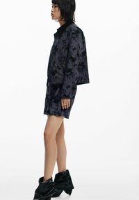 Jachetă întunecată, cu un model, și un set scurt asortat, realizat dintr-un material texturat. Prezintă un guler și cizme negre supradimensionate distinctive.