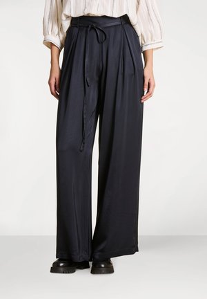 Personne portant un pantalon noir large à taille haute avec une fine ceinture, associé à des chaussures noires épaisses et un chemisier de couleur claire à manches 3/4.