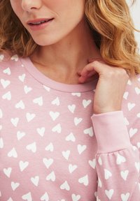 Maglia a maniche lunghe di colore rosa chiaro con un motivo testurizzato di piccoli cuori bianchi, colletto e polsini a coste, e tessuto morbido.