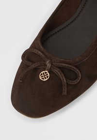 Καφέ μπαλαρίνα από σουέντ με στρογγυλεμένη μύτη, διακοσμημένη με φιόγκο και μεταλλικό γούρι. Η μαλακή εσωτερική επένδυση προσφέρει άνε ση.