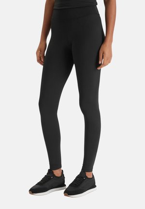 Persona che indossa leggings neri a vita alta e scarpe da ginnastica nere, in piedi su uno sfondo chiaro e uniforme.