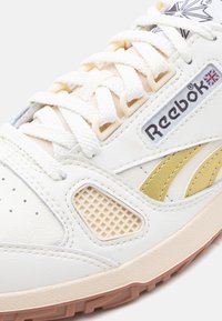 Λευκό και μπεζ αθλητικό παπούτσι Reebok με υφή στην σόλα, λευκά κορδόνια, χρυσές λεπτομέρειες στο πλάι και τη σημαία του Ηνωμένου Βασιλείου κοντά στο λογότυπο της μάρκας.