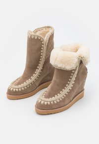 Botas de cuña de ante marrón con un suave forro de piel de peluche y costuras decorativas a lo largo de los bordes, con una suela plana de goma para mayor tracción.