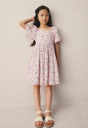 Young girl standing with hand on head, wearing a pink floral dress with puff sleeves and beige sandals against a plain background.