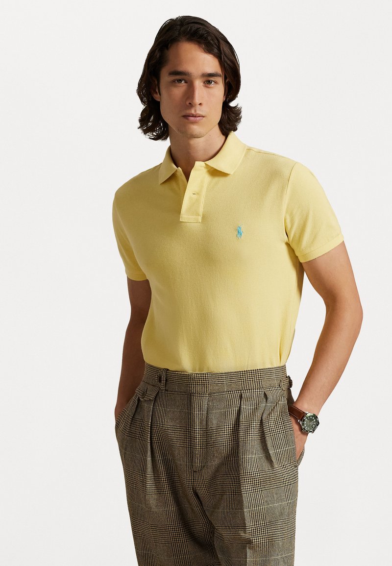 Dzeltena polo krekls ar īsām piedurknēm un apkakli, ar mazu tirkīza logotipu. Valkāts kopā ar raibiem biksēm un pulksteni.