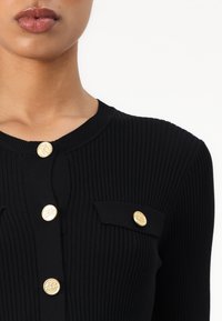 Cardigan noir à côtes avec boutons ronds dorés et deux petites poches à rabat. Design ajusté avec un col rond.